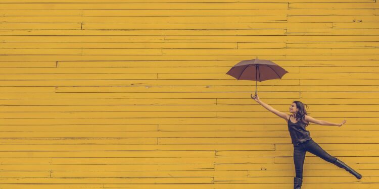 tanzende Frau mit Regenschirm vor gelber Wand