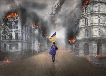 Kleiner Junge mit Fahne der Ukraine geht durch eine Straße mit brennenden Wohnhäusern