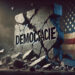 Dreidimensionaler Schriftzug "Democracie" auf bzw. in einer bröckelnden Wand, daneben die US-Amerikanische Flagge, AI generiert