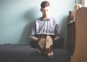 Junger Mann sitzt in einem vermutlich Studentenzimmer auf dem Bett und liest ein Buch, Symbolbild für "Musterschüler"