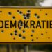 Ortseingangsschild mit der Aufschrift "DEMOKRATIE" und zahlreichen Einschußlöchern, AI generiert