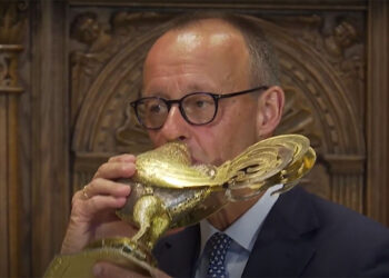 Merz trinkt aus einer goldenen Weinkanne