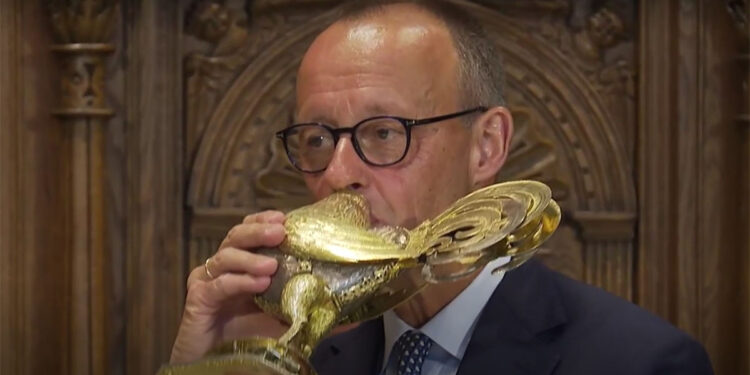 Merz trinkt aus einer goldenen Weinkanne