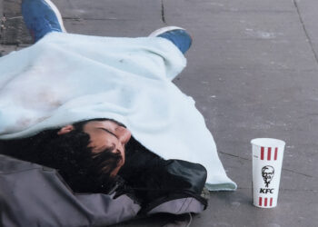 Obdachlose Person schläft auf der Straße, neben ihm ist ein Becher für Spenden aufgestellt