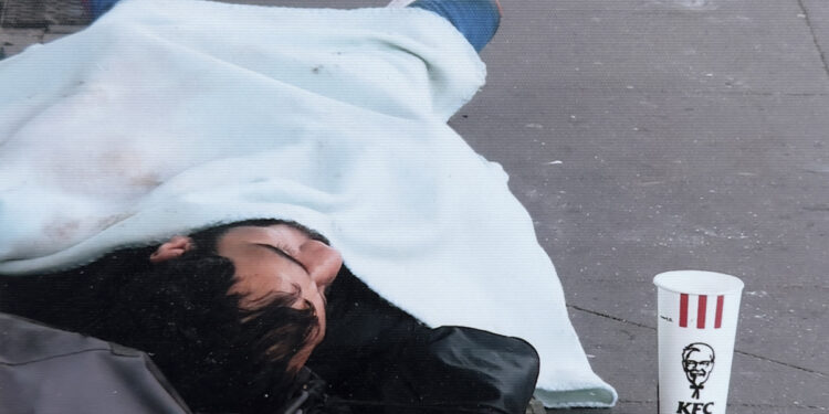 Obdachlose Person schläft auf der Straße, neben ihm ist ein Becher für Spenden aufgestellt