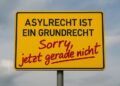 Ortseingangschilc "Asylrecht ist ein Grundrecht" mit Sprayzusatz "Sorry, jetzt gerade nicht". AI generiert