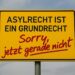 Ortseingangschilc "Asylrecht ist ein Grundrecht" mit Sprayzusatz "Sorry, jetzt gerade nicht". AI generiert