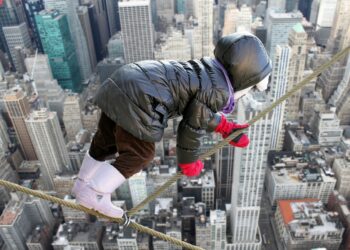 KInd in Winterkleidung mit roten Handschuhe klettert in schwindelnder Höhe auf einem Seil über einem abgrund vor der Kulisse einer Stadt mit Wolkenkratzern