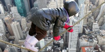 KInd in Winterkleidung mit roten Handschuhe klettert in schwindelnder Höhe auf einem Seil über einem abgrund vor der Kulisse einer Stadt mit Wolkenkratzern